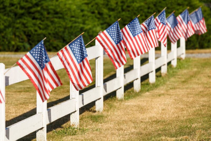 Patriotic display of American flags waving on white picket fence. Typical small town Americana Fourth of July Independence Day decorations.